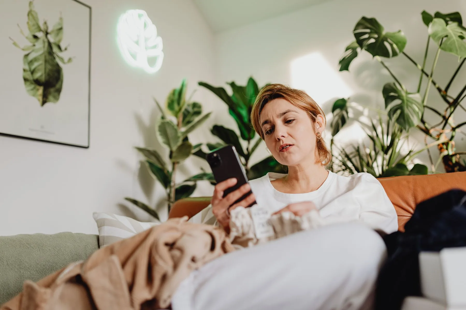 A woman sitting on her sofa looking at her phone, surrounded by houseplants including a Monstera.